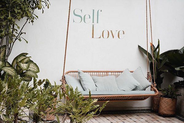 On this image there is a garden with a bench. Behind the bench, there is a bench which says self-love. This image represents the topic self-care love language well.