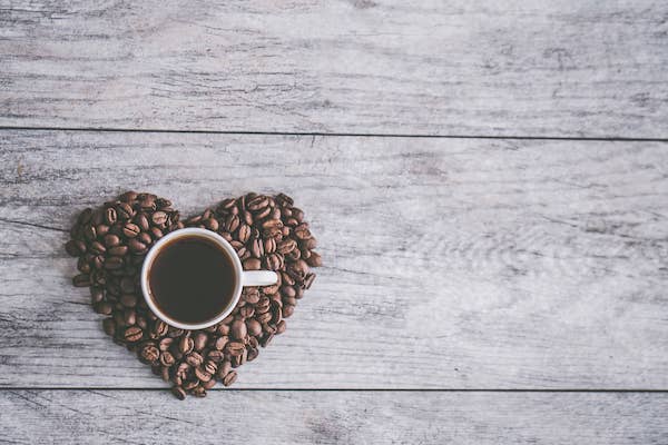 One of the signs that you are a highly sensitive person is that you can't handle caffeine well. On this image, there is a cup of coffee with some coffee beans shaped in the form of a heart under it.