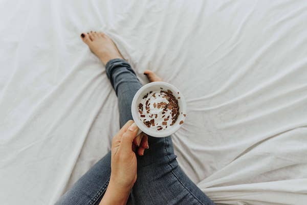 Spend less time on your phone by going offline. On the image, there is a woman sitting on the bed with a cup of hot chocolate, enjoying doing nothing.
