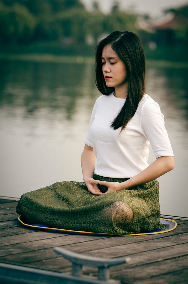 On the image, there is a woman sitting on a deck meditating. She is covered with a blanket to keep herself warm.
