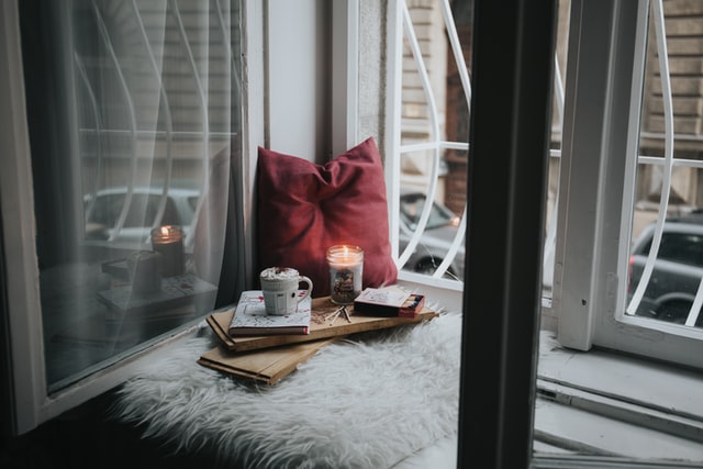 On the picture, there is a window nook with a piece of faux fur on it. On the faux fur, there is a pillow with a candle in front of it. Also, there is a book and a cup of hot chocolate.