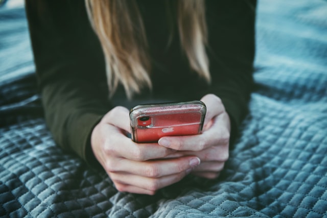 On the image, there is a woman looking at her phone. She is lying on her bed.