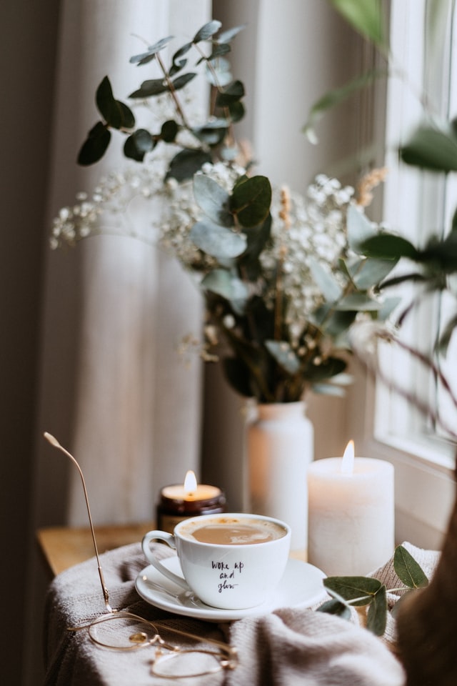 On the image there is a window with a cup of coffee in front of it, a few candles, a pair of glasses and a flower bouquet in the background.