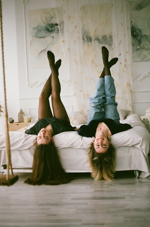 On the image, there are to women who stay calm because they are spending time together and laughing. They are lying on a bed with their head over the edge of the bed and they are smiling. It looks like they are friends.
