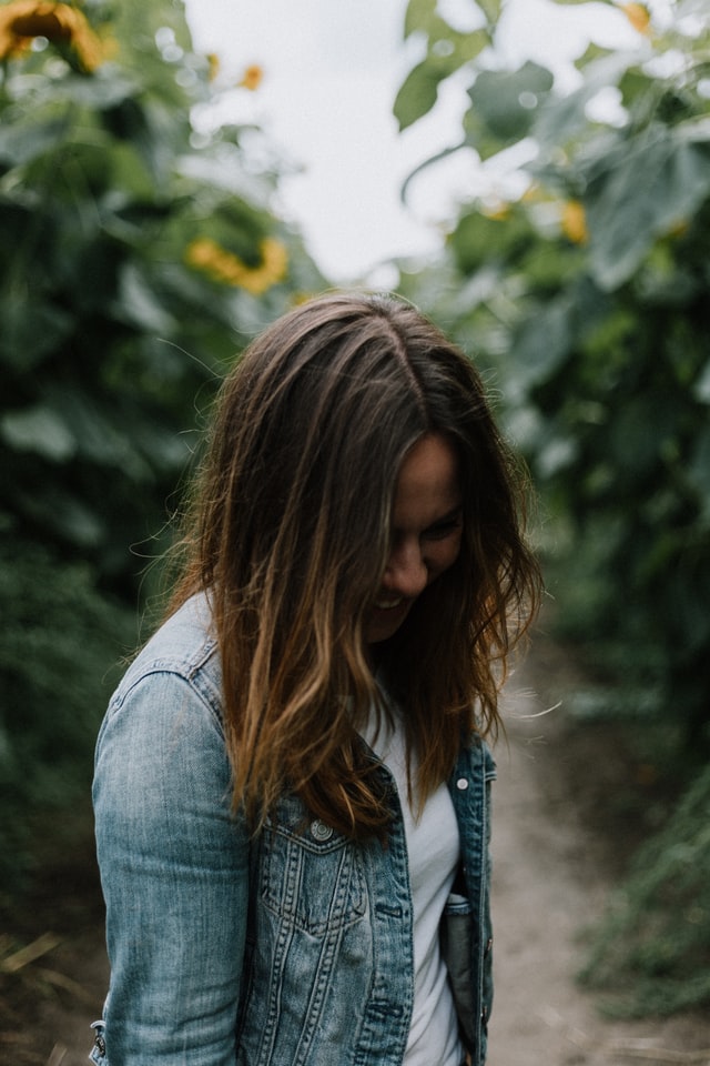 On the image, there is a caucasian woman standing in a sunflower field. She is looking down and smiling. She is wearing a jeans jacket and a white tshirt. 