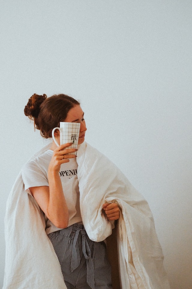 On the image, there is a young caucasian woman with a blanket wrapped around her. She is holding a cup of tea in front of her face. She looks somewhat tired, like she is in need of self-love and inner peace.
