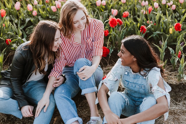 On the image, there are three young woman laughing. They are sitting in front of a field of flowers.