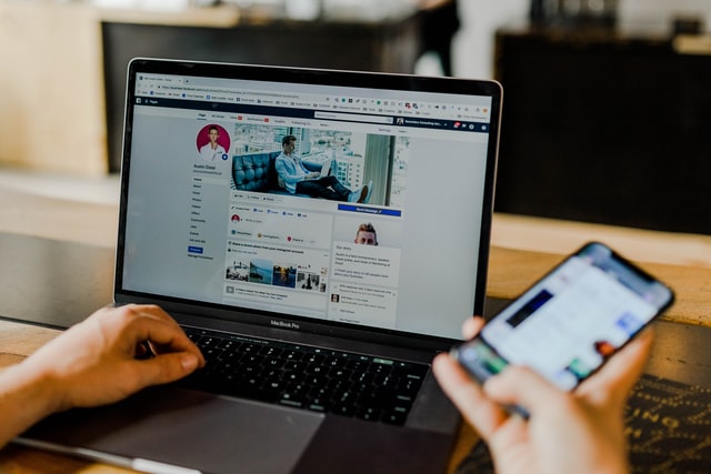 On the image, there is a laptop and we see two hands. One hand is located on the keyboard of the laptop, while the other is holding a mobile phone. On both screens, a social media app is opened. The photo represents social boundaries for the introvert perfectly.