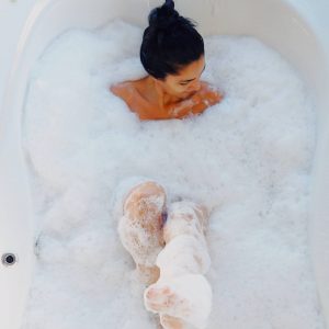 A Highly Sensitive woman enjoying a relaxing bath filled with puffy white bubbles, with only her head and legs poking out of the frothy bubbles. Photo Credit: Matheus Frade