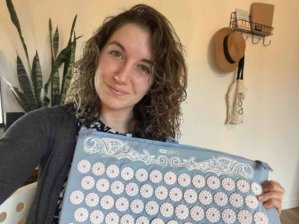 A woman with curls holding the shakti mat in her hands and smiling at the camera.